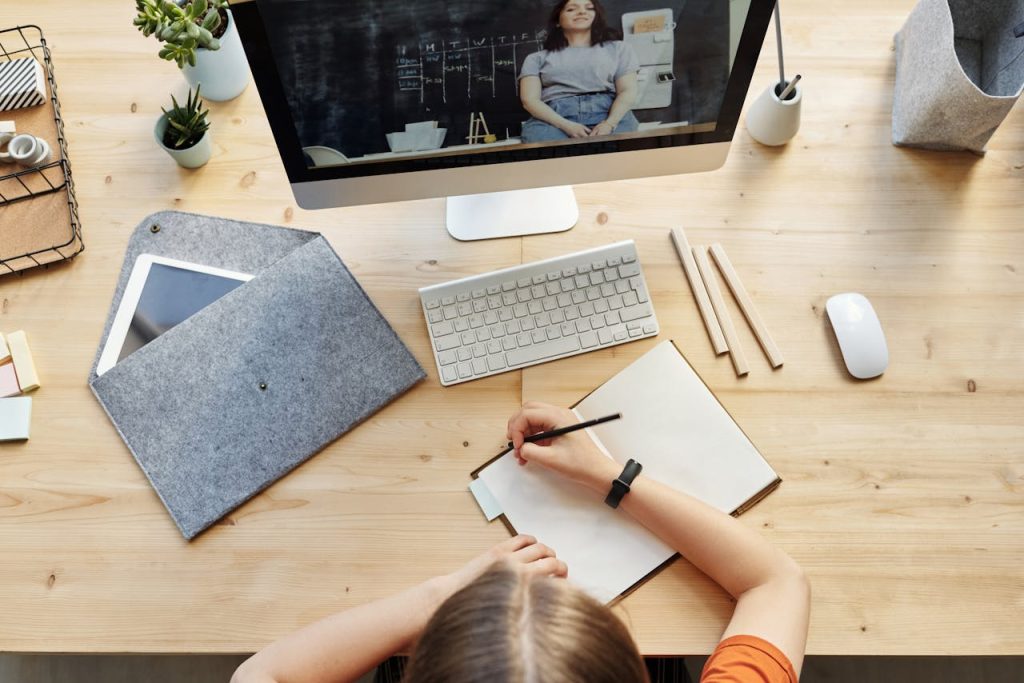 A teenager studying from home using a computer and notebook in a well-organized workspace.